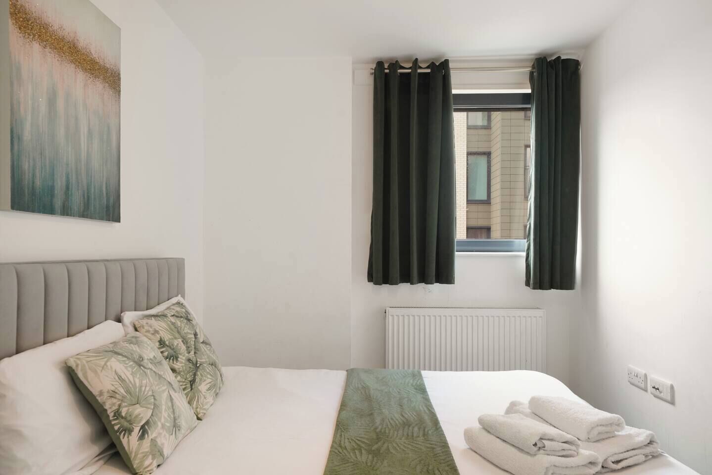 A hotel room with a bed, a window with curtains, a radiator, and a painting on the wall.