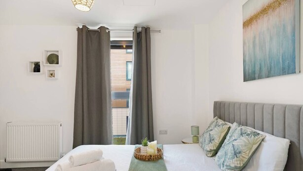 Una camera d'albergo con un letto, un termosifone, una finestra con tende, un comodino con una lampada e un quadro appeso al muro.