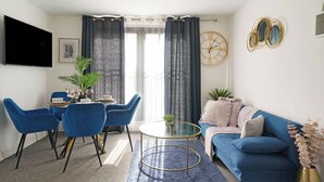 Un saló modern amb un sofà blau, una taula de dinar amb cadires, un televisor fixat a la paret i una finestra gran amb cortines.