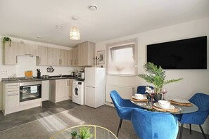 A modern kitchen with built-in appliances, a dining area with blue chairs, and a large flat-screen TV mounted on the wall. - Heathrow Gateway Apartments with Private Parking35 (Harlington)