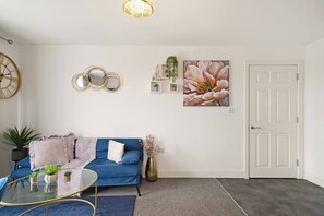 A living room with a blue sofa, a glass coffee table, and a white door. - Heathrow Gateway Apartments with Private Parking35 (Harlington)