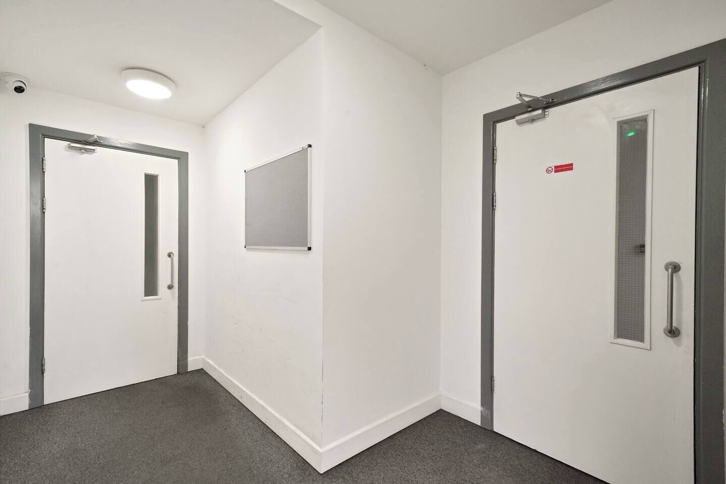 A corridor with white walls, grey carpet, and doors with metal handles and a red sign.