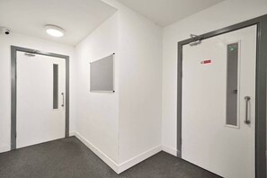 A corridor with white walls, grey carpet, and doors with metal handles and a red sign. - Heathrow Gateway Apartments with Private Parking13 (London)