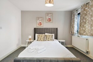 A neatly made bed with pillows, a bedside table with a lamp, and a radiator. - Heathrow Gateway Apartments with Private Parking13 (London)