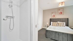 A hotel room with a shower, a bed with a grey headboard, and two wall-mounted artworks. - Heathrow Gateway Apartments with Private Parking13 (London)