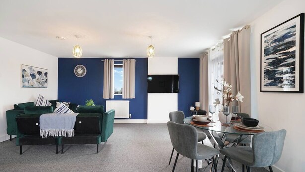 A modern living room with a green sofa, a dining table with chairs, a flat-screen TV, and artwork on the walls. - Heathrow Gateway Apartments with Private Parking13 (London)
