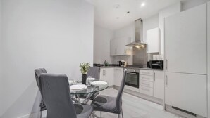 A modern kitchen with white cabinetry, a glass dining table, and grey chairs. - Stylish 1BR Near Heathrow & Elizabeth Line 8 (London)