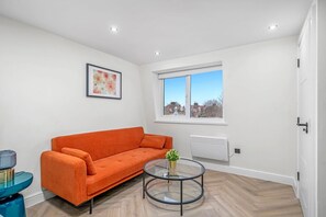 A modern living room with a sofa, a glass coffee table, a radiator, and a window with a view.