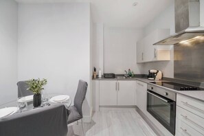 A compact kitchen with a dining area, featuring a modern stove, white cabinets, and a small table with chairs. - Stylish 1BR Near Heathrow & Elizabeth Line 8 (London)