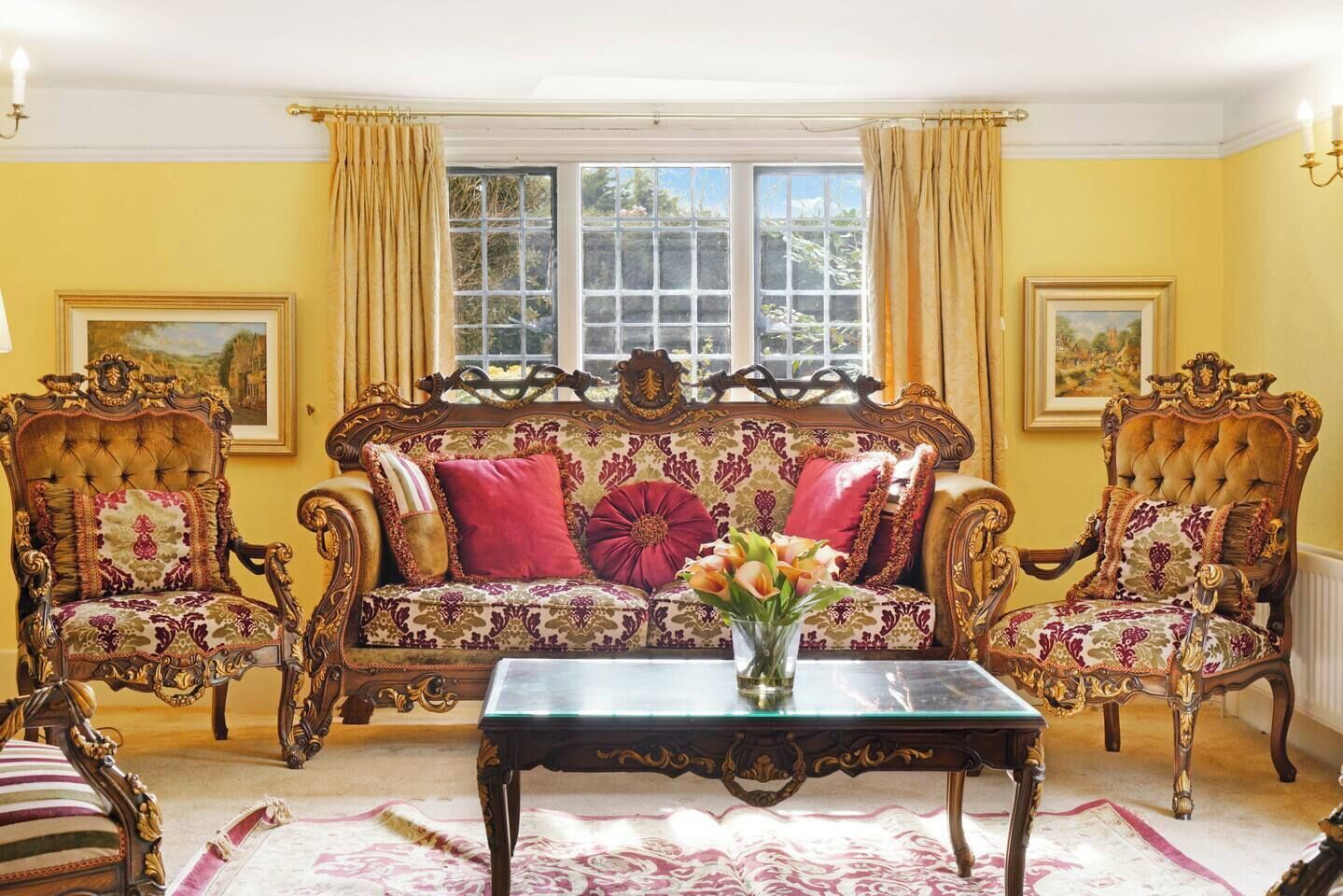Et værelse med en stor, detaljeret sofa, to ens matchende lænestole, et glasklædt sofabord og en vase med blomster.