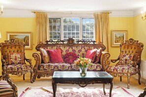 Ein Zimmer mit einem großen, verzierten Sofa, zwei passende Sessel, einem Glastisch und einer Blumenvase.