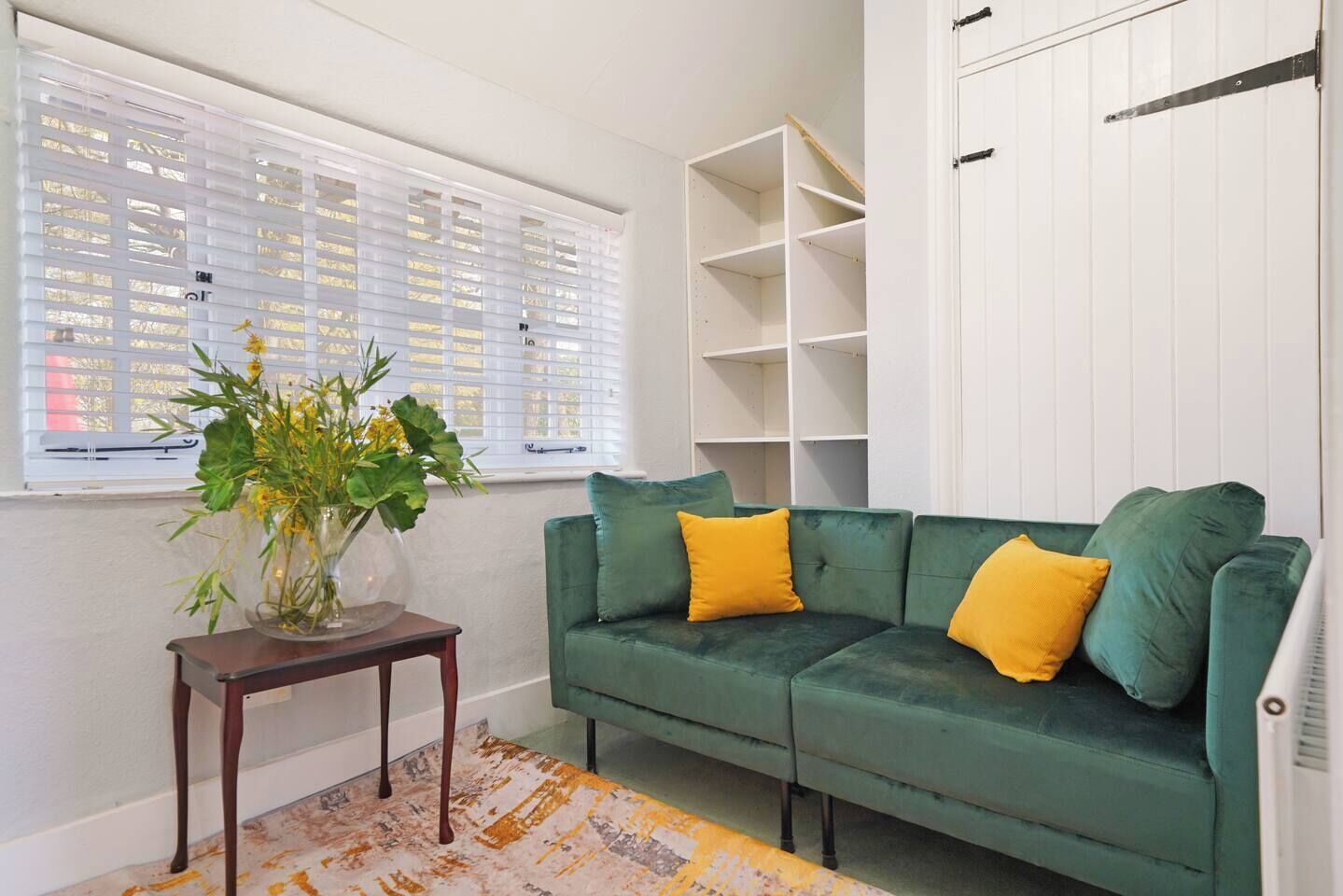 A green sofa with yellow cushions, a wooden side table with a vase of flowers, and a white shelving unit against a white wall.