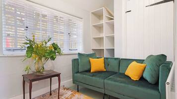 A green sofa with yellow cushions, a wooden side table with a vase of flowers, and a white shelving unit against a white wall.