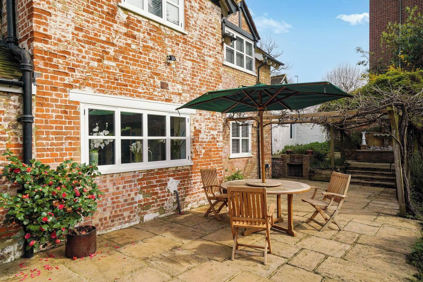 A brick house with a patio area featuring a round wooden table and chairs, a green umbrella, and a potted plant with red flowers.