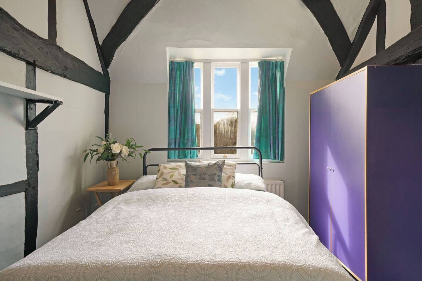 A bedroom with a bed, a window with curtains, a bedside table with a vase, and a wardrobe.