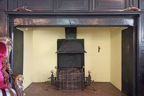 A fireplace with a metal grate, flanked by two ornate metal stands, set in a room with a yellow wall and a dark wooden mantel. - 16th Century manor house | 6BR | Private Grounds (Harlington)