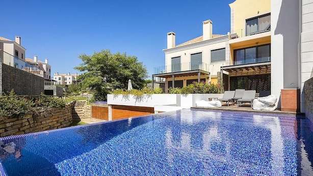 Un área de piscina moderna con una piscina azul cristalina, paredes blancas y un balcón con sillones de descanso.