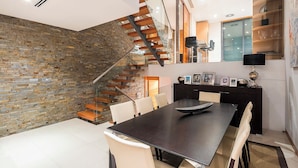 Un comedor moderno con una mesa de madera oscura, sillas blancas, una escalera de cristal y una pared de piedra.