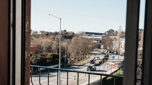Vista da janela com corrimão de metal, céu claro e uma estrada com carros e edifícios ao longe.
