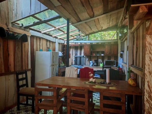 A rustic wooden dining area with a table and chairs, a refrigerator, and a kitchen in the background. - DondeLALO Hospedaje (Vara Blanca)