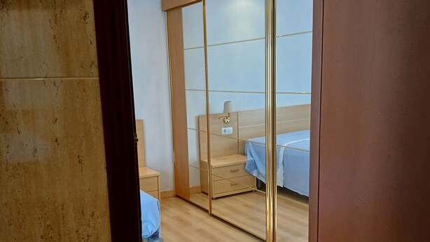 Ett modernt hotellrum med en stor skjutdörr i glas, en säng med ett blått överkast, ett sängbord och ett litet bord vid sängen.