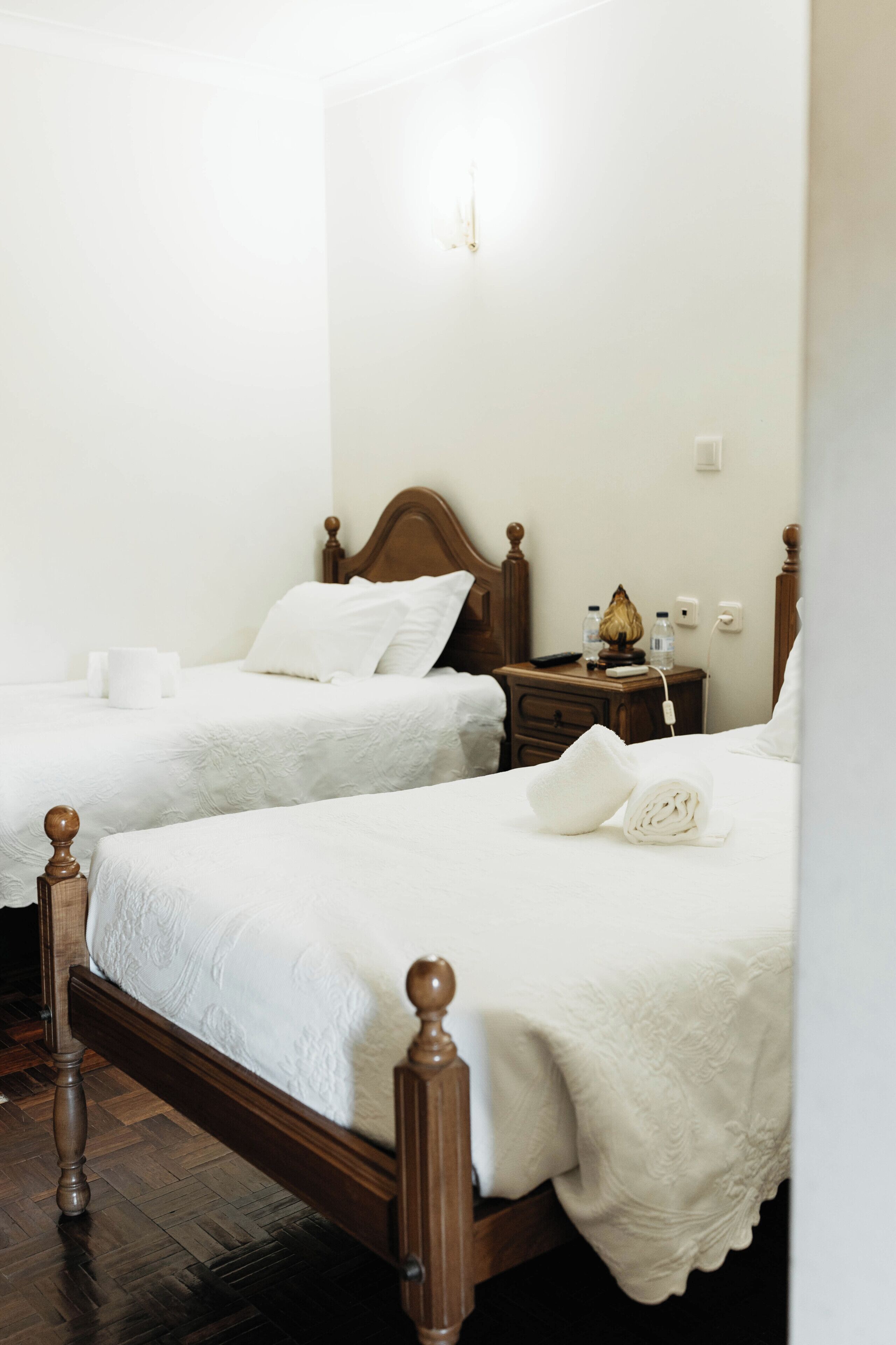 A hotel room with two wooden beds, white linens, a bedside table with a lamp, and a power outlet.