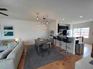 Eine moderne Küche mit Barbereich, einem Esstisch mit Stühlen und einem grauen Sofa in einem Wohnbereich mit Deckenventilator und Pendelleuchten.