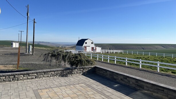 A white barn with a dark roof, a white fence, and a paved area with stone edging. - Century Farm in Horse Heaven Hills, Tri-Cities WA (Kennewick)