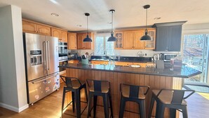 Een moderne keuken met houten kasten, roestvrijstalen apparatuur, een centraal eiland met barkrukken, hanglampen en uitzicht op de omgeving.