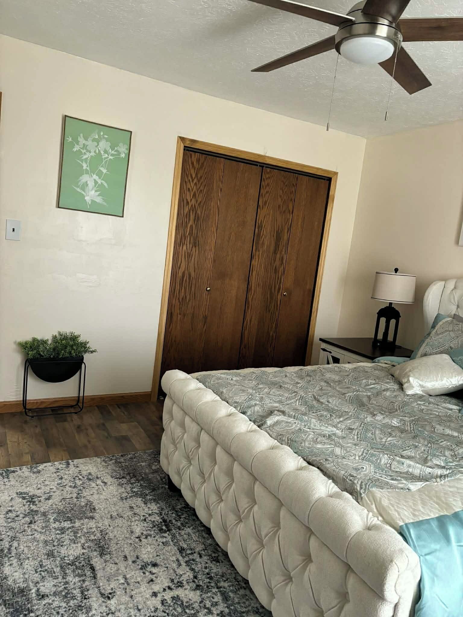 Camera da letto con un letto, una ventola a soffitto, un armadio in legno, un tavolino con una lampada e una pianta in vaso.