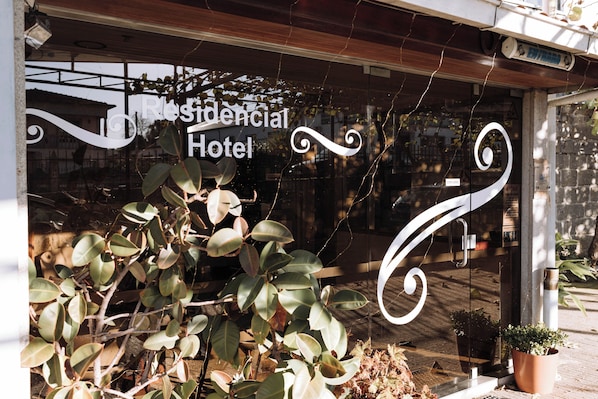 Entrada de un hotel con un letrero que dice 'Residencial Hotel', diseños de vidrio decorativos y plantas en macetas.