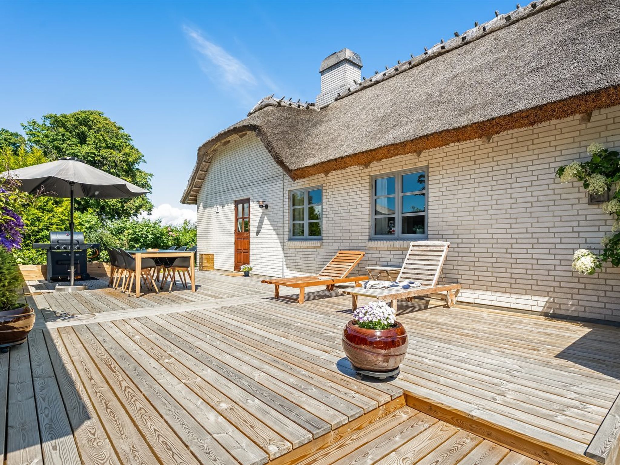 Ett hus med halmtak, en träveranda, utomhusmöbler och en grillplats.