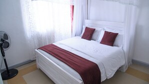 A bed with white bedding and red pillows, a standing fan, and lace curtains. - LA FONT HOUSE Near JKIA Nairobi (Nairobi)