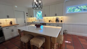 Cocina con armarios blancos, una isla central con taburetes, una lámpara colgante y un ventanal con vista a árboles.
