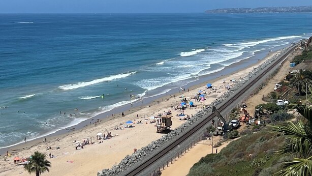 Una playa con una extensa hilera de sillas y sombrillas, una torre de salvavidas y una vía de tren que corre paralela a la playa.