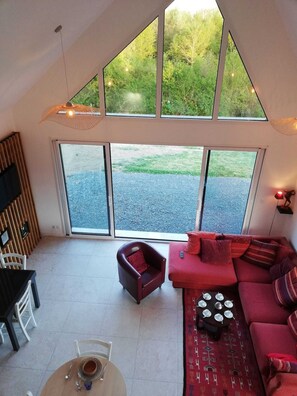 A modern interior with a red sofa, a small table, and a chair, large windows offering a view of greenery.