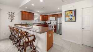 Una cocina moderna con armarios de madera, una isla central con taburetes y electrodomésticos de acero inoxidable, incluyendo un refrigerador y un microondas.