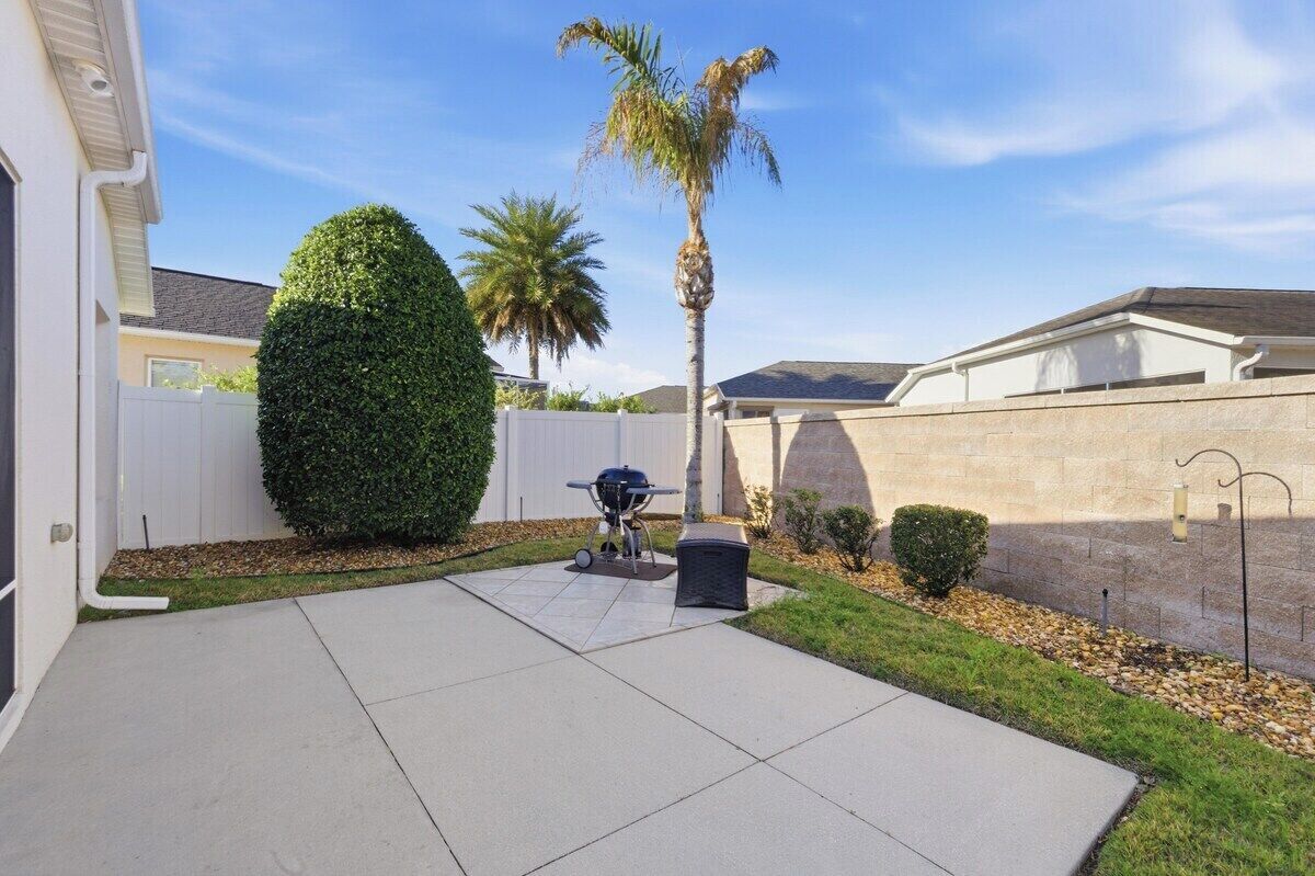 A residential backyard with a patio, a barbecue grill, a palm tree, and a well-maintained hedge.