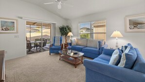 Un salón con un sofá azul, una mesa de centro de vidrio, un ventilador de techo y vistas a un patio exterior a través de puertas corredizas de cristal.