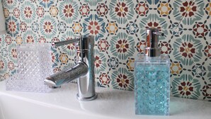 A bathroom with a patterned tile backsplash, a chrome faucet, and two clear glass containers on the sink. - Room 8 A (Trapani)