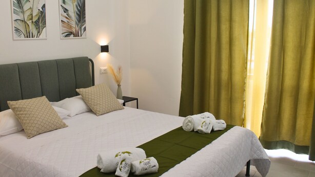 A neatly made bed with white linens and green bedspread, a bedside table with a lamp, and two framed botanical prints on the wall. - Room 8 A (Trapani)