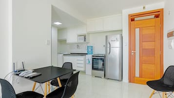 A compact kitchen with white cabinets, a microwave, a refrigerator, and a water dispenser. There is a dining table with chairs and a wooden door.