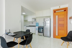 Eine kompakte Küche mit weißen Schränken, einer Mikrowelle, einem Kühlschrank und einem Wasserspender. Ein Esstisch mit Stühlen und eine Holztür sind ebenfalls vorhanden.