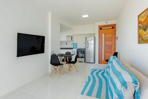 Ein Wohnzimmer mit einer Couch, einem an der Wand befestigten Fernseher, einem Essbereich mit Stühlen und Tisch sowie einer Küche mit Geräten.