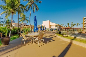 An outdoor patio area with tables and chairs, surrounded by palm trees and a clear blue sky. - VT08 Great Apt for 3 people - Beachfront Cond. (Lauro de Freitas)