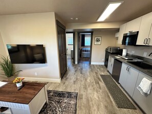 A modern kitchen with a flat-screen TV, a dining area with a wooden table, and a hallway leading to a bedroom. - Private Ptarmigan Suite (Hayden)