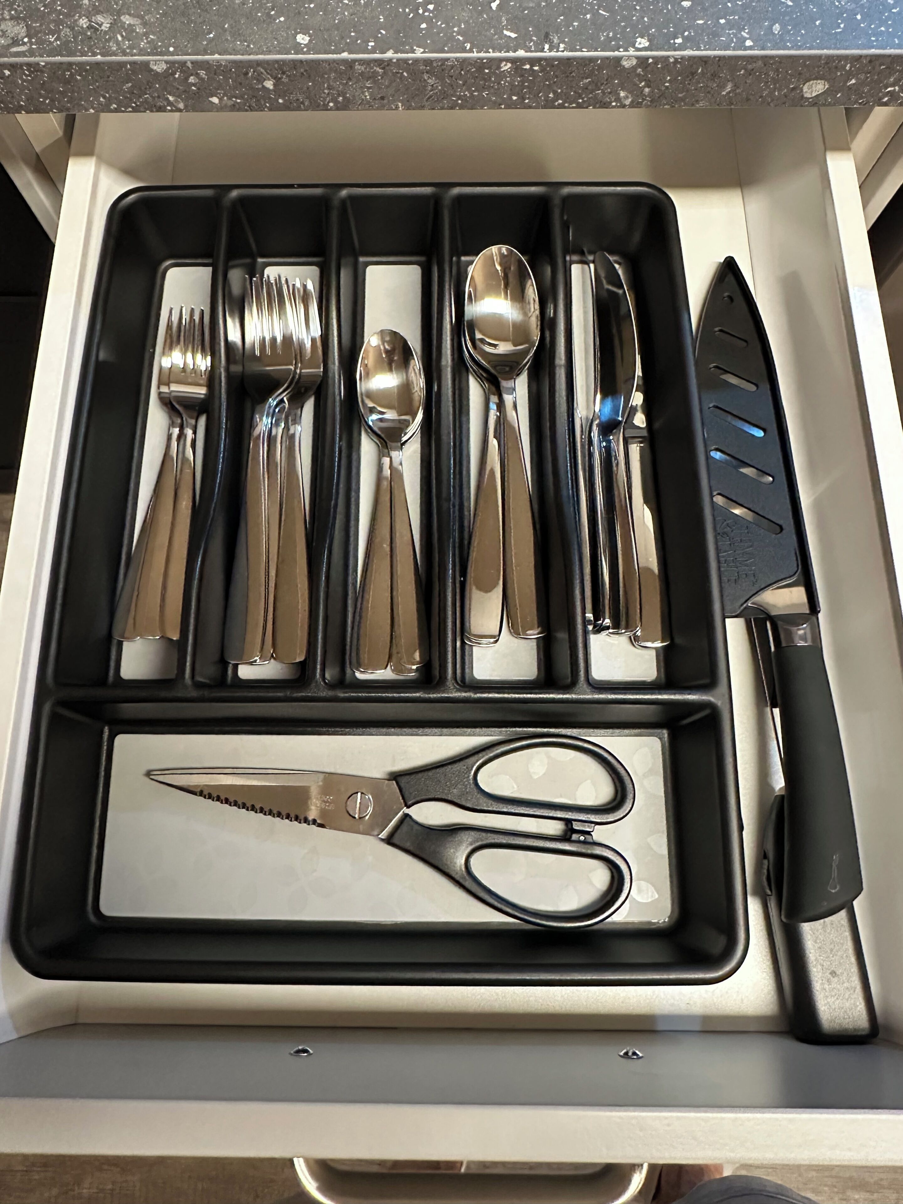A drawer containing a set of silverware including forks, spoons, and a knife, along with a pair of scissors.