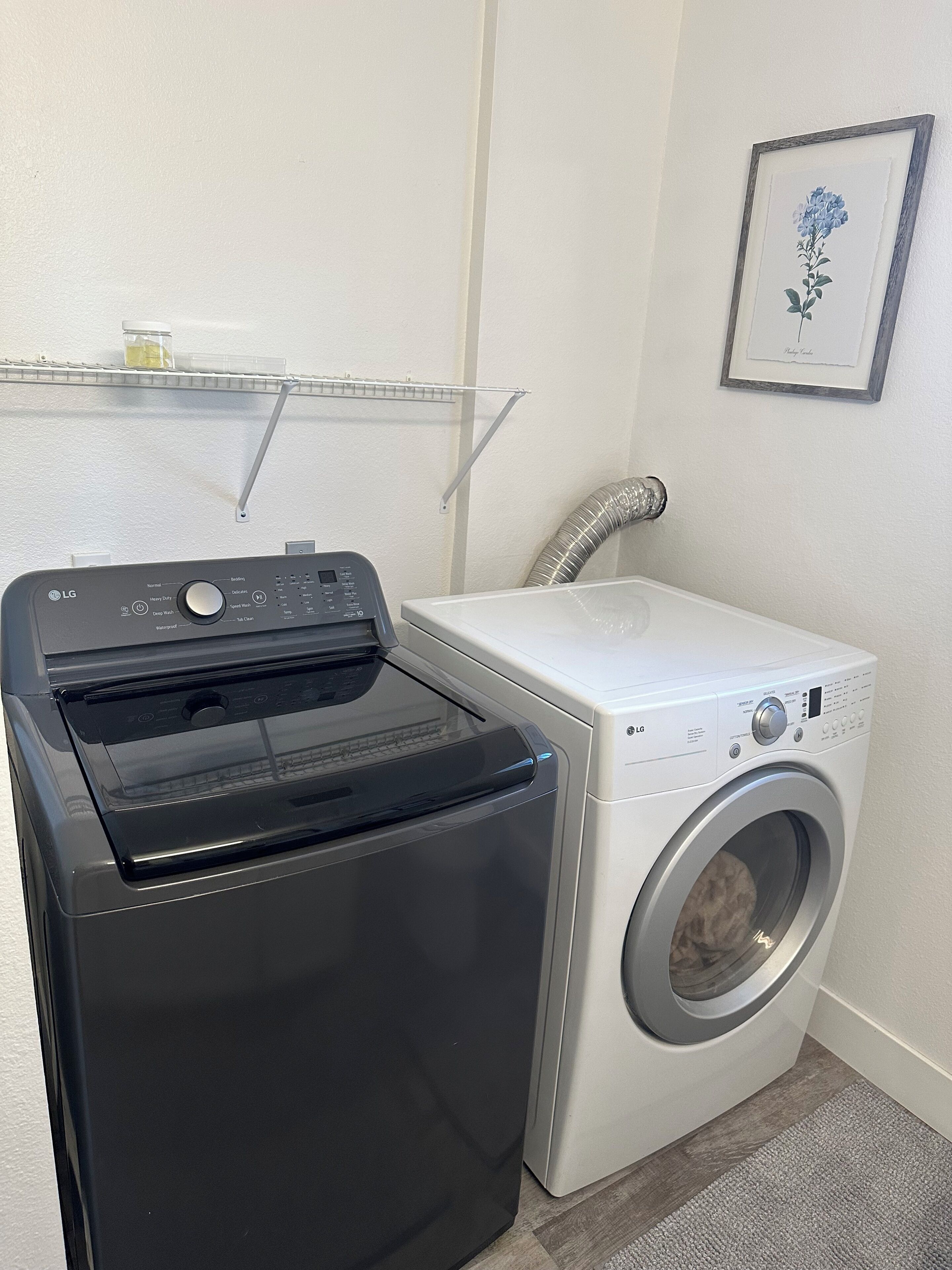 A laundry area with a black dryer, a white washing machine, a metal shelf with a jar, and a framed picture on the wall.