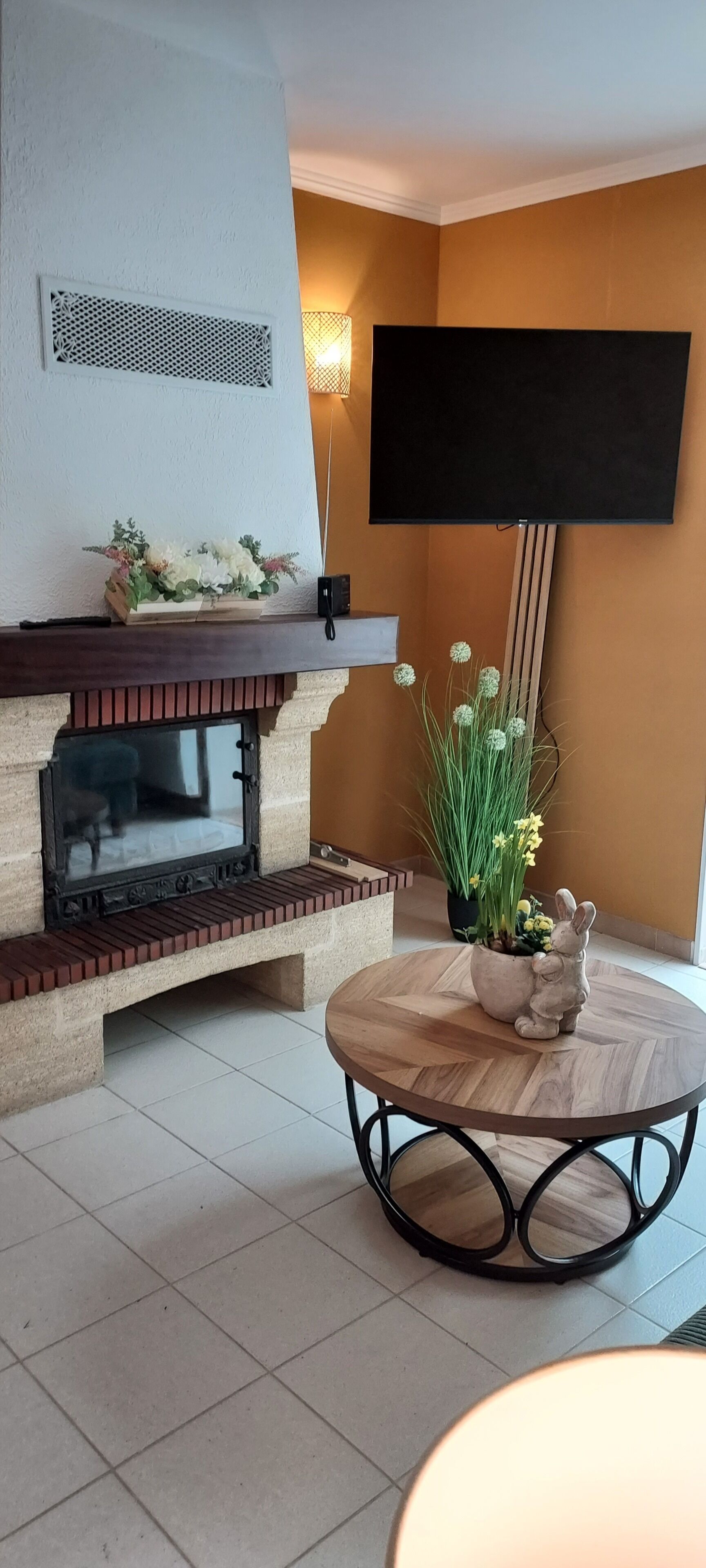 Een woonkamer met een open haard, een flatscreentelevisie aan de muur, een ronde houten tafel met metalen poten en een bloeiende plant in pot.