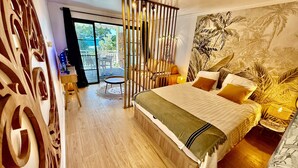 Ein Schlafzimmer mit einem Bett, einem kleinen runden Tisch, einem Sessel, einer Wand mit Dekoration und Blick durch eine Glastür.